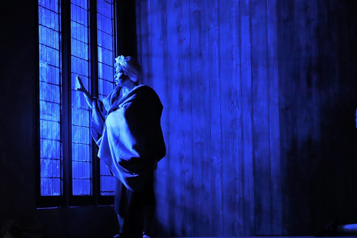 An actress in a headwrap and shawl stands at a moonlit window in a staged production